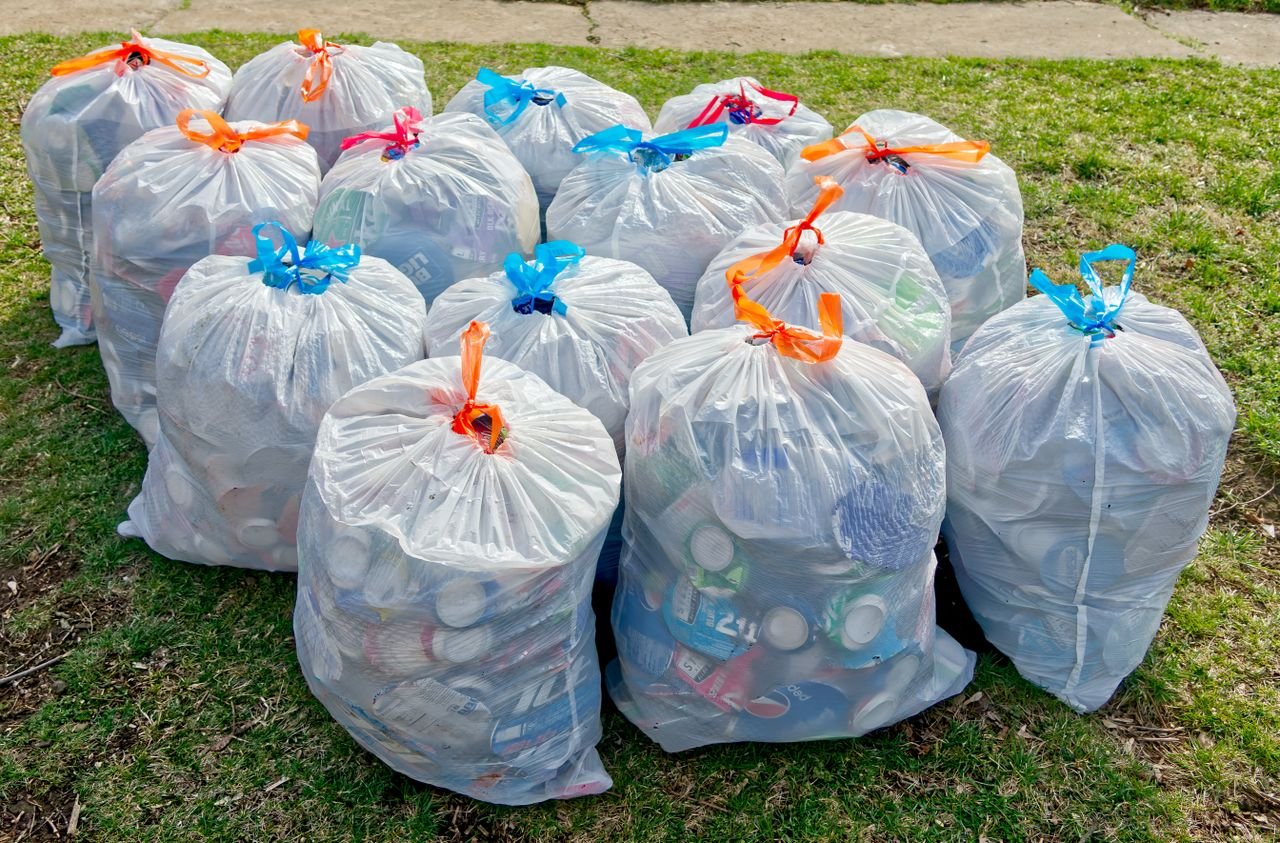 who-we-are Plastic trash bags filled with recyclable materials on grass, raising recycling awareness.