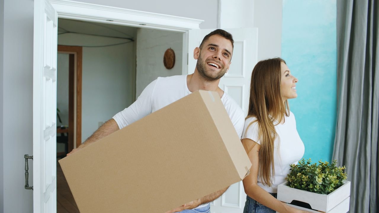 services-02 A cheerful couple moving into their new home, carrying boxes and a plant.