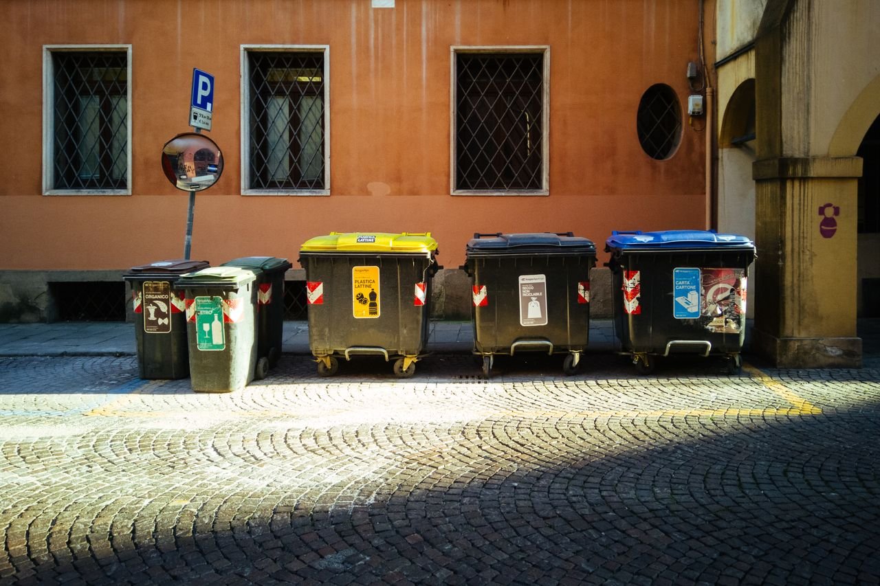 our-story Five recycling bins on a sunny cobblestone street against an orange building.